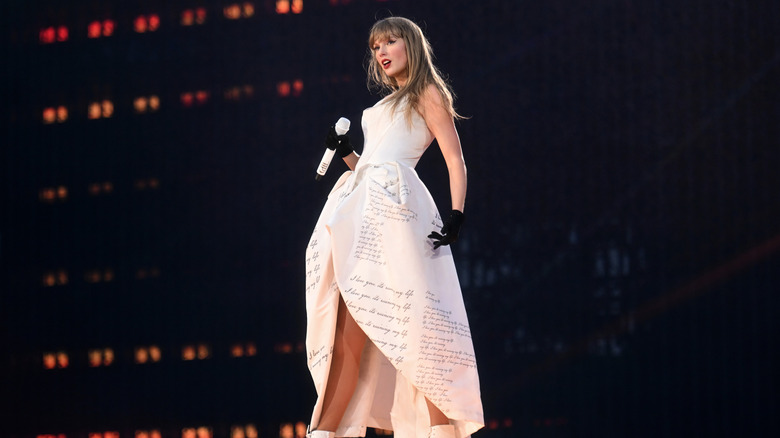 Taylor Swift posa en el escenario con un vestido blanco y guantes negros, mientras sostiene un micrófono en una mano.