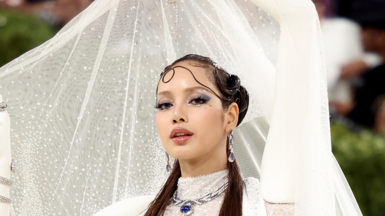 Lisa de Blackpink posando en la Met Gala 2026 con un vestido blanco con parte del mismo flotando detrás de su rostro.