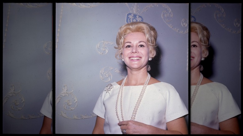 Eva Gabor sonríe luciendo un collar de perlas y un vestido blanco contra una pared azul.