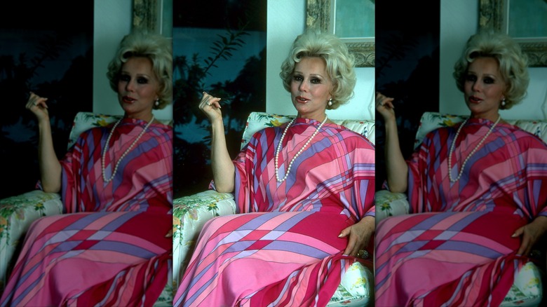 Eva Gabor posa con un vestido rosa, morado y azul sobre un sofá con la mano en alto.
