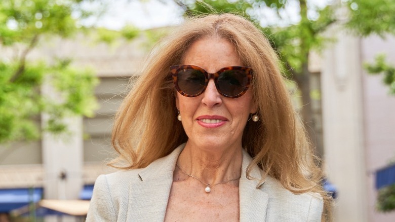 Una mujer de pelo largo que viste una chaqueta color canela, joyas de perlas y gafas de sol.