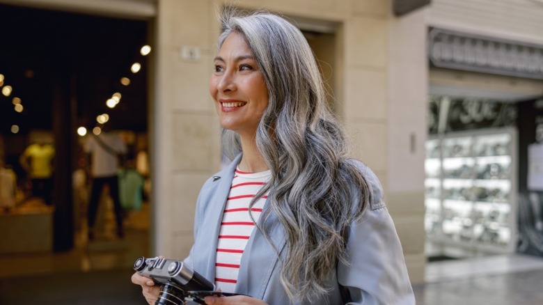 Una mujer con cabello largo y ondulado sonriendo y sosteniendo una cámara, vestida con una camisa a rayas rojas y blancas y una chaqueta gris.