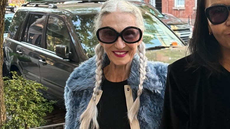 Una mujer con dos largas trenzas sonriendo en la calle con una chaqueta peluda y grandes gafas de sol.