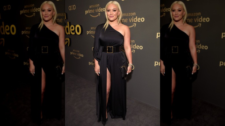 Hilary Duff sonriendo en un evento de alfombra roja luciendo un vestido negro de un solo hombro con un gran cinturón.