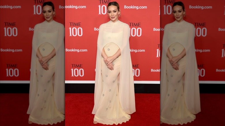 Hilary Duff posando con las manos delante en un evento de alfombra roja con un vestido color crema con capa.