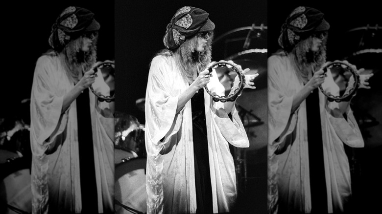Fotografía en blanco y negro de Stevie Nicks con un sombrero flexible y una bata blanca tocando una pandereta actuando con Fleetwood Mac en 1977.