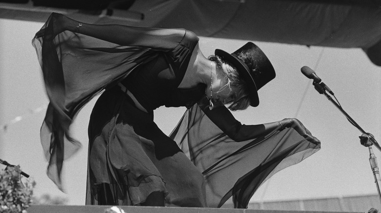 Fotografía en blanco y negro de Stevie Nicks vistiendo un traje negro fluido con un sombrero de copa negro en el escenario actuando con Fleetwood Mac en 1977.
