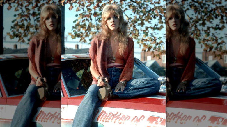 Stevie Nicks posa en un auto con jeans y una blusa roja con zapatos de plataforma y flequillo de cortina.
