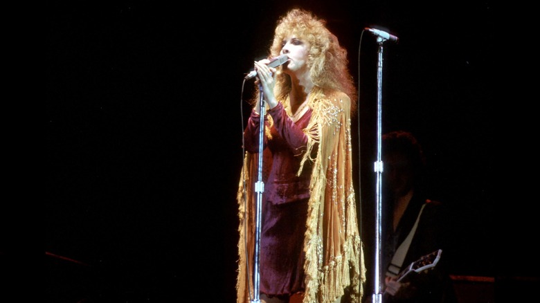 Stevie Nicks vistiendo un traje rojo con una capa dorada cantando frente a un micrófono actuando con Fleetwood Mac en 1977.