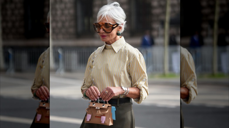 Una mujer de pelo corto y gris que viste una camisa con cuello a rayas y gafas de sol con anillos dorados y plateados y una pulsera plateada.