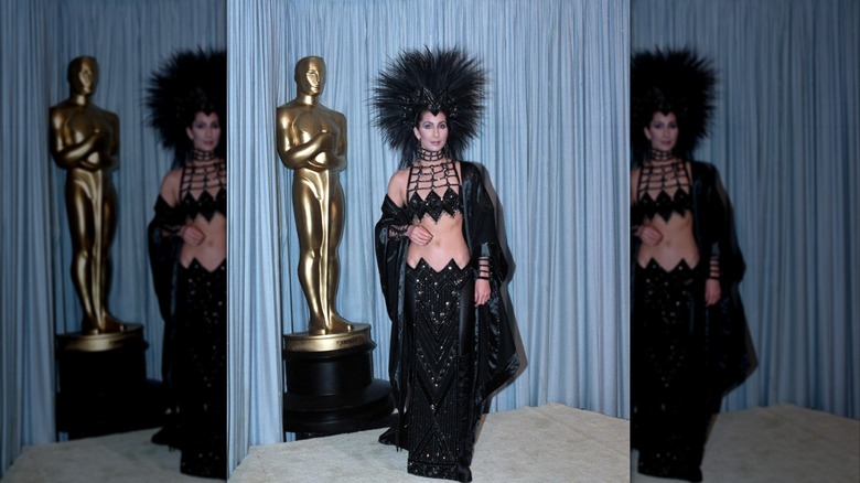 Cher con un traje negro brillante y un gran tocado negro de plumas frente a una estatua del premio en los Oscar de 1986.