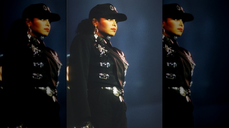 Janet Jackson posando en el escenario con una gorra negra y un traje negro de inspiración militar durante su gira Rhythm Nation.