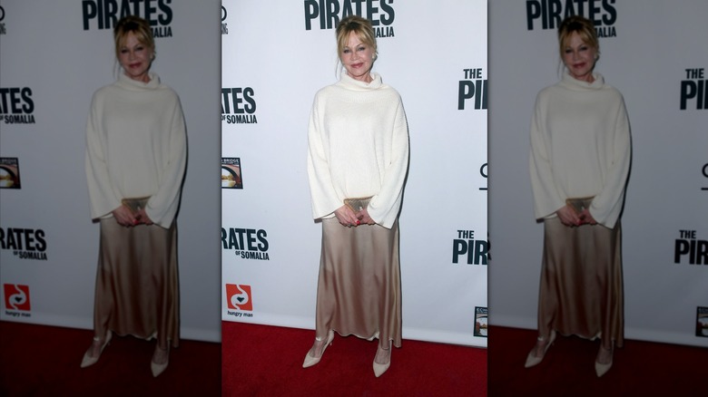 Melanie Griffith sonríe luciendo un jersey de cuello alto blanco y una falda de satén beige.