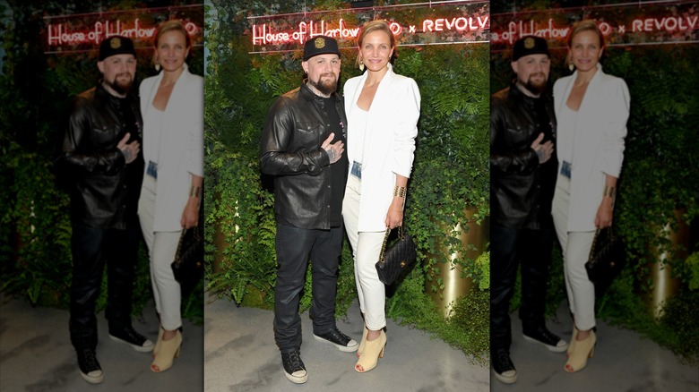 Benji Madden vistiendo un traje completamente negro y un sombrero posando frente a un fondo de hojas con Cameron Diaz vistiendo un look completamente blanco.
