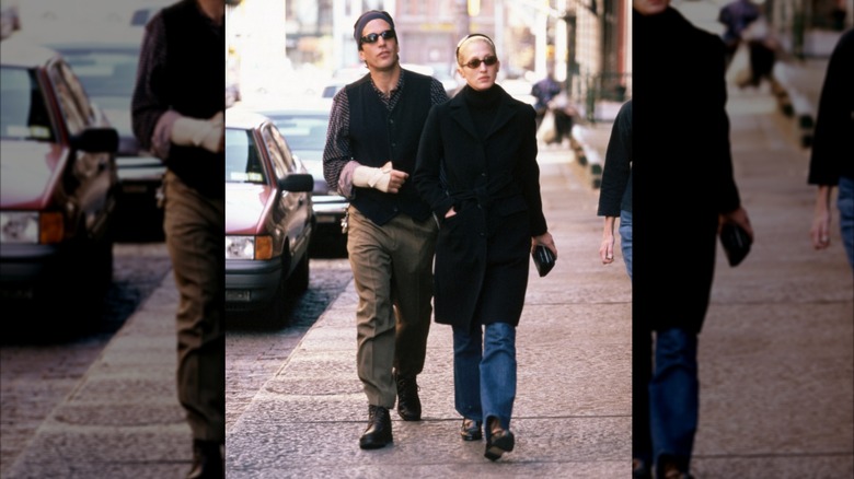 Carolyn Bessette-Kennedy con jeans bootcut, gabardina negra y botas de tacón caminando junto a JFK Jr.