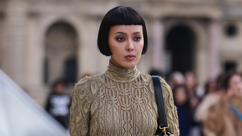 Kiwi Lee Han viste cabello negro con un corte de pelo bob con flequillo, un suéter de cuello alto de punto beige, una minifalda a cuadros a cuadros negra y roja, un gran cinturón amarillo y verde con patrones de rombos, un bolso de cuero negro, botines de cuero negro adornados con pedrería, afuera de Louis Vuitton, durante la Semana de la Moda de París - Ropa de mujer Otoño/Invierno 2026/2027, el 10 de marzo de 2026
