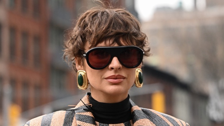 Una mujer con gafas de sol, pendientes grandes y el pelo ondulado con un corte de duendecillo.