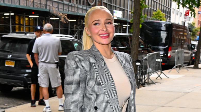 Hayden Panettiere caminando afuera con el cabello recogido en una cola de caballo y una chaqueta a cuadros blanca y negra.
