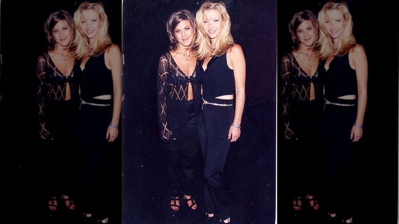 Lisa Kudrow con pantalones negros y un mini chaleco negro posando junto a Jennifer Aniston.