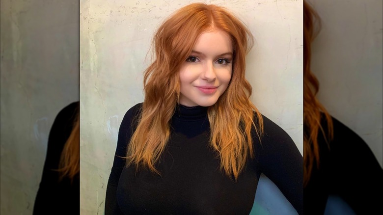 Ariel Winter posando con un cuello de tortuga negro y cabello rojo ondulado sobre un fondo claro.