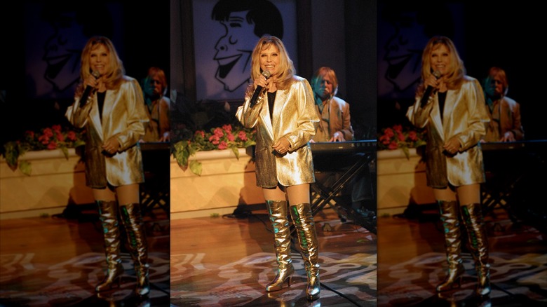 Nancy Sinatra actuando en el escenario sosteniendo un micrófono con botas doradas hasta la rodilla y una chaqueta dorada.