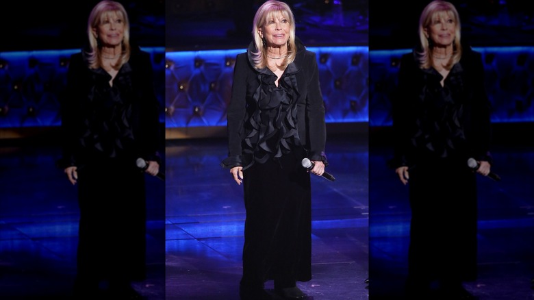 Nancy Sinatra sosteniendo un micrófono en el escenario con un top negro con volantes y una falda negra.
