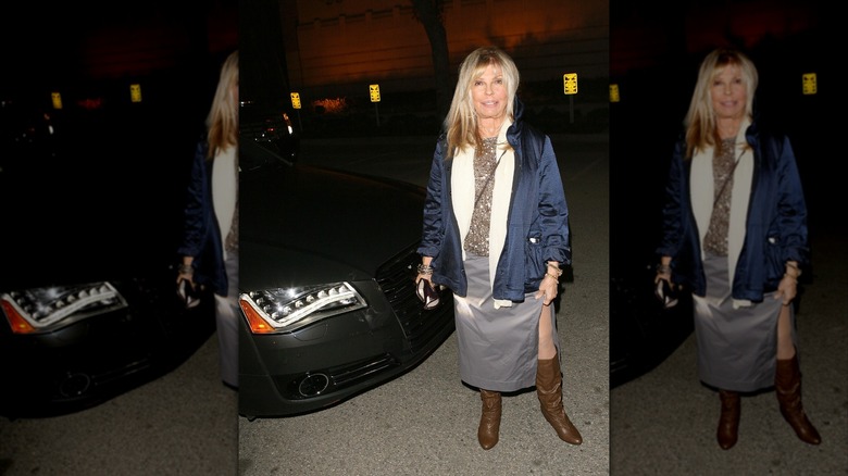 Nancy Sinatra posando en un evento con una falda plateada y un top dorado brillante con un abrigo azul oscuro y botas marrones.