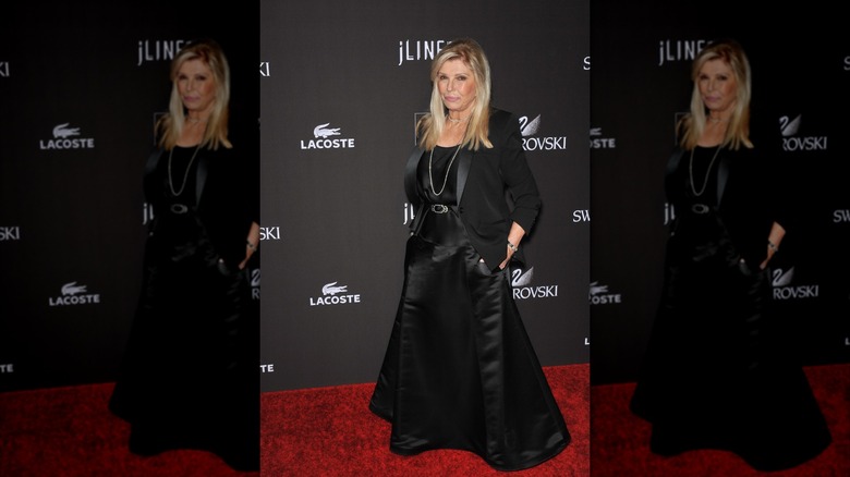 Nancy Sinatra posando en un evento de alfombra roja con las manos en los bolsillos luciendo una falda de seda negra y una chaqueta negra.
