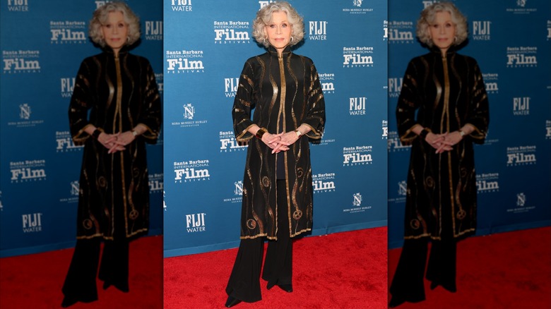 Jane Fonda posando en un evento de alfombra roja con pantalones negros fluidos y una túnica negra y dorada.
