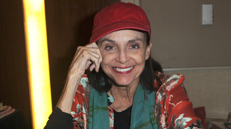 Valerie Harper sentada en una mesa sonriendo y con un sombrero rojo.