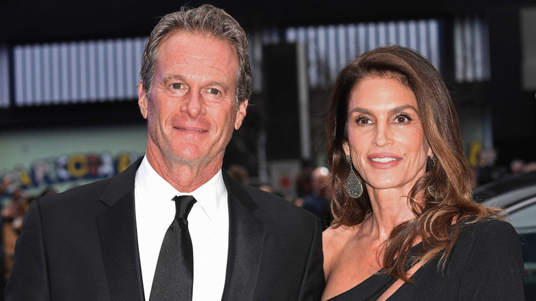 Rande Gerber con un traje negro al lado y Cindy Crawford con un vestido negro posando afuera.