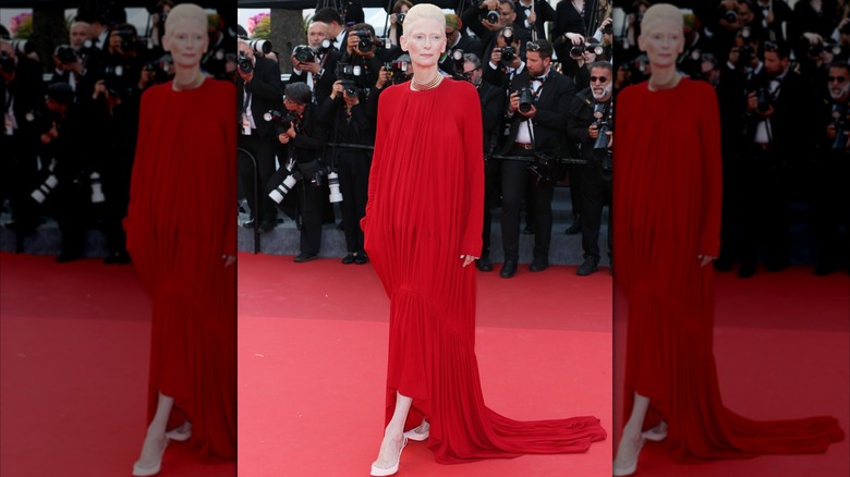 Tilda Swinton posa en la alfombra roja con el pie en punta y mirando hacia un lado con un vestido rojo fluido.