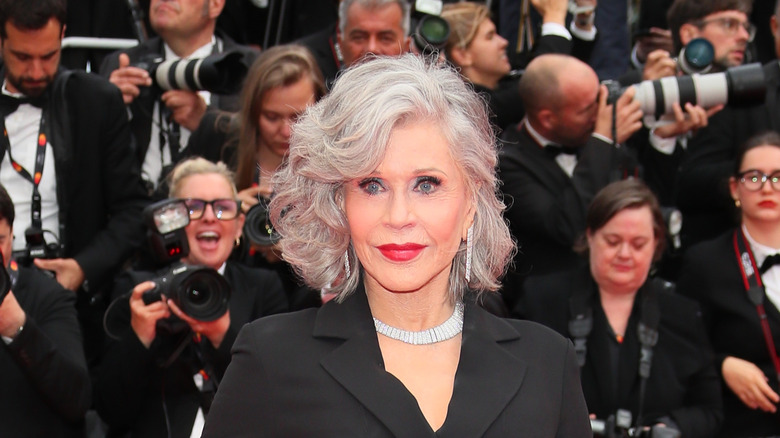 Jane Fonda posando en un evento de alfombra roja luciendo lápiz labial rojo, una chaqueta negra y un collar de diamantes.
