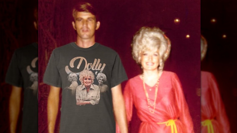 Dolly Parton sonríe mientras toma de la mano a su esposo Carl Dean, quien está retocado con Photoshop para que use una de sus camisetas comerciales.