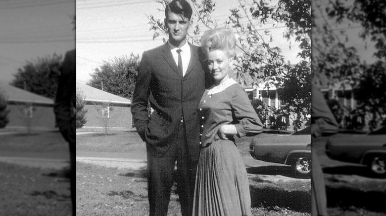 Una fotografía en blanco y negro de Carl Dean y Dolly Parton posando juntos afuera frente a un auto antiguo.
