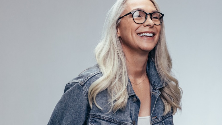 Una mujer sonriente con el pelo largo y gris, con gafas y una chaqueta vaquera.