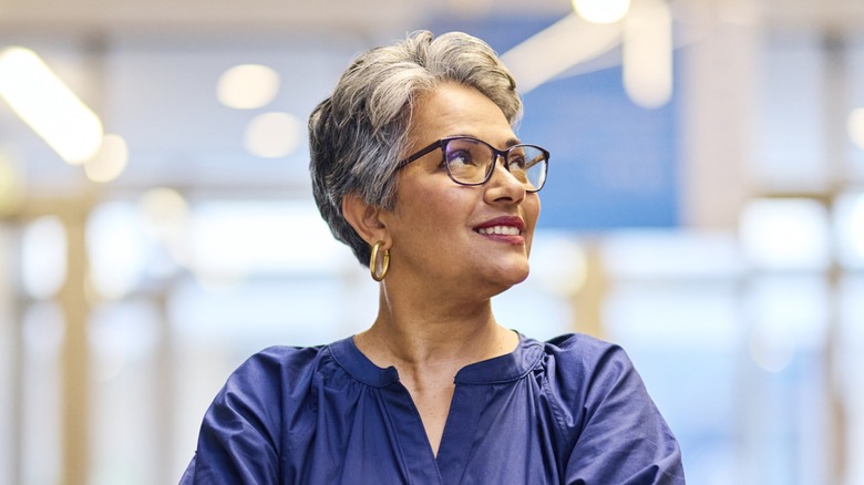 Una mujer con un corte pixie de color gris oscuro, con gafas y un top azul, mirando hacia un lado.