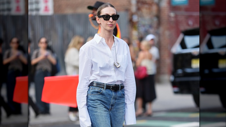 Una mujer con camisa blanca, jeans y un collar plateado con un gran colgante.