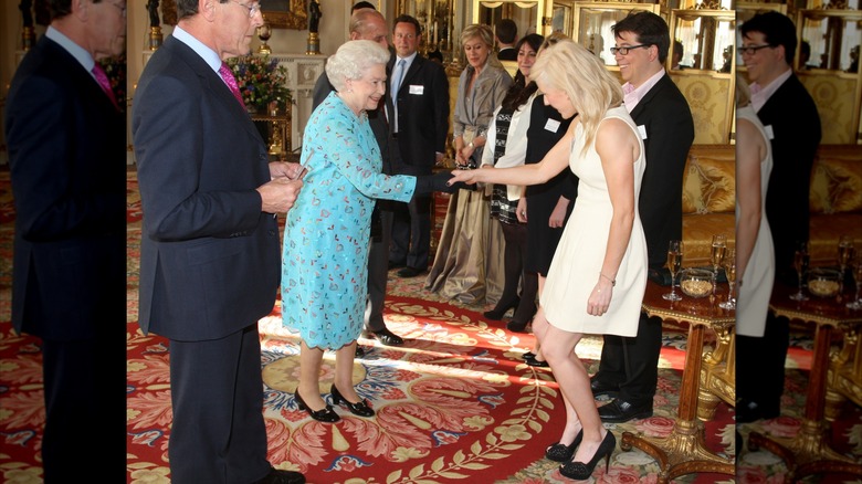 Ellie Goulding con un vestido blanco y zapatos negros haciendo una reverencia y estrechando la mano de la reina Isabel con un vestido azul.