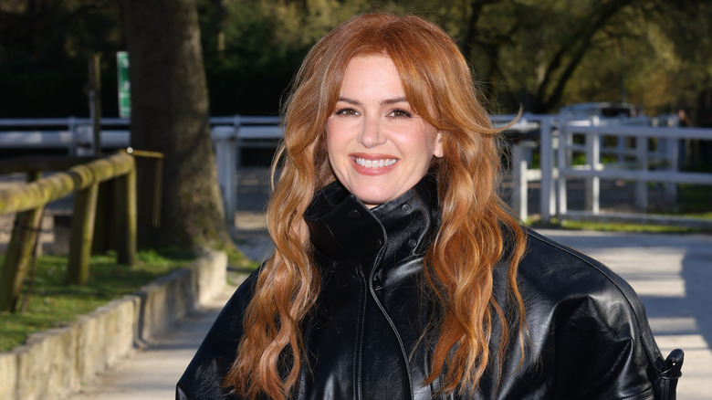 Isla Fisher sonriendo afuera con una chaqueta de cuero negra y el cabello suelto.