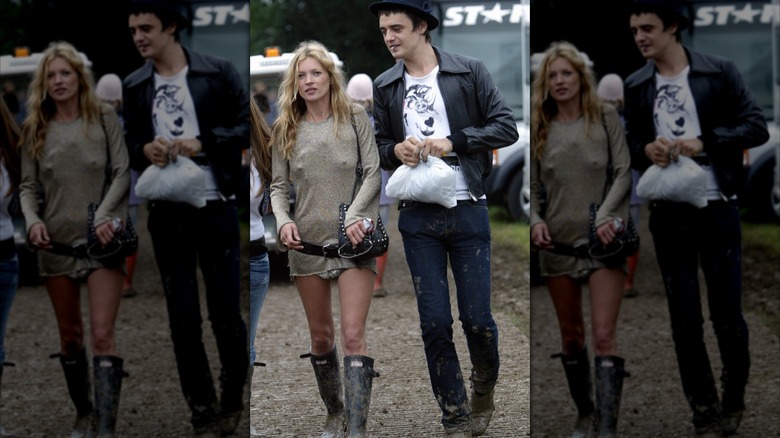 Kate Moss con un atuendo desaliñado con botas altas y Pete Doherty con jeans, camisa blanca y chaqueta bomber negra, caminando afuera.