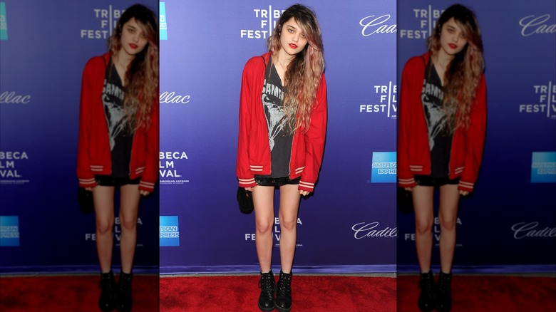 Sky Ferreira posando en un evento de alfombra roja con la cabeza hacia un lado y vistiendo una chaqueta roja, camiseta de la banda, pantalones cortos negros y botas de combate.
