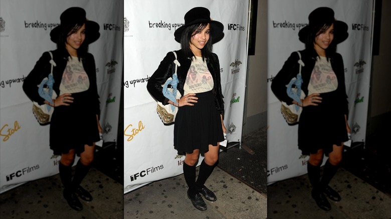 Zoë Kravitz posa en el estreno de una película con la mano en la cadera, vestida con una chaqueta y falda negras, una camiseta estampada blanca y un sombrero negro de ala ancha.