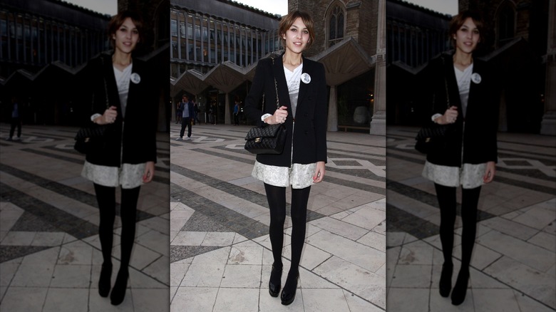 Alexa Chung fotografiada con una chaqueta negra, vestido blanco, medias negras y zapatos negros al aire libre.