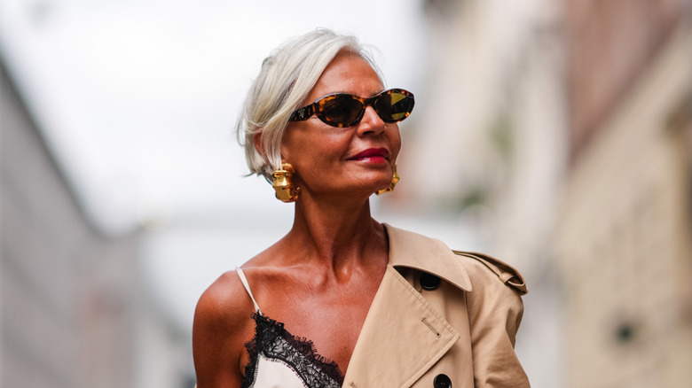 Una mujer posa con gafas de sol ovaladas y una gabardina beige que le cuelga del hombro.