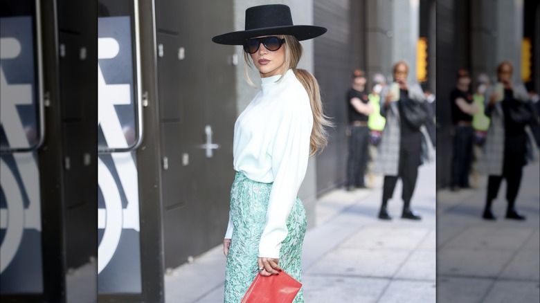 Jennifer López posando afuera en Nueva York con un sombrero negro de ala grande con gafas de sol, un top blanco y una falda verde.