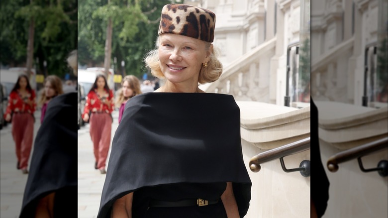 Pamela Anderson sonriendo afuera con un vestido caplet negro y un sombrero tipo pastillero con estampado de leopardo.