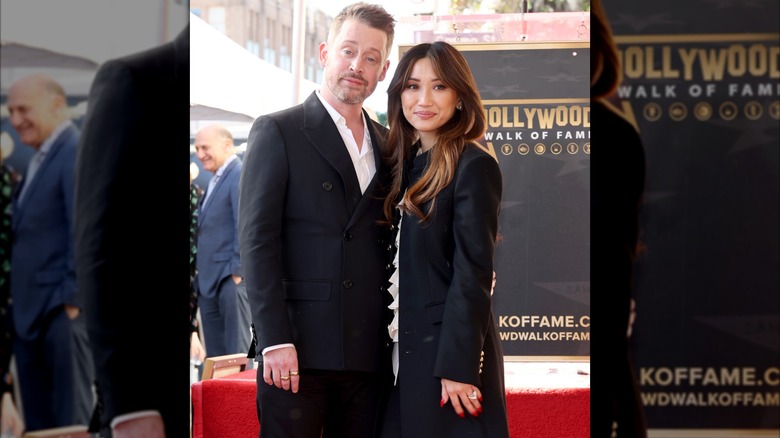 Macaulay Culkin con un traje negro junto a Brenda Song con una gabardina larga negra.