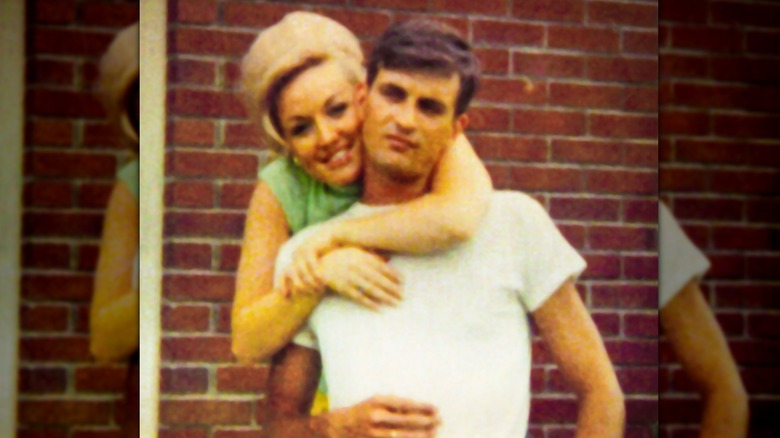 Una joven Dolly Parton aparece fotografiada frente a una pared de ladrillos mientras abraza a su esposo Carl Dean por detrás.