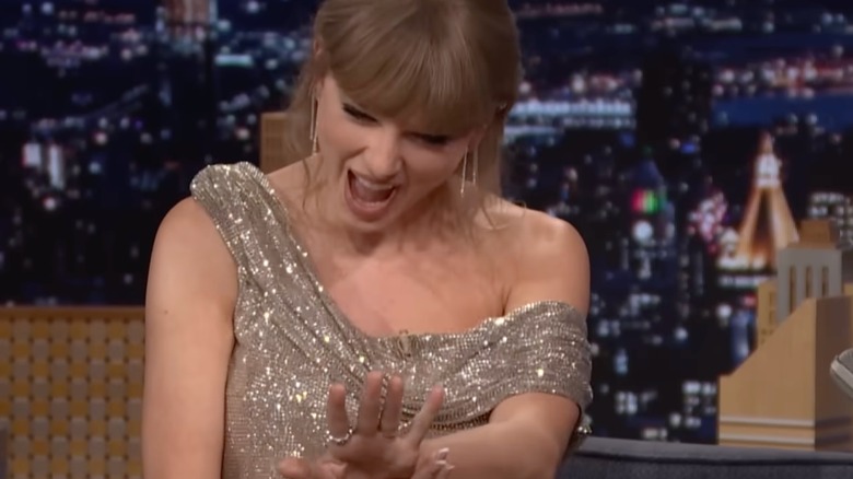 Taylor Swift sonríe mientras mira su anillo de compromiso durante una aparición televisiva nocturna.